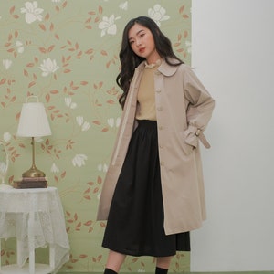 K&ouml;nnte beinhalten: Ein beiger Trenchcoat mit einem wei&szlig;en Streifen an den &Auml;rmeln und einem schwarzen Rock. Der Mantel hat einen Kragen mit wei&szlig;em Besatz und einen G&uuml;rtel in der Taille. Das Model tr&auml;gt schwarze Socken und schwarze Schuhe.