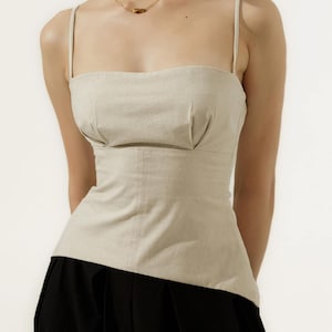 May include: A beige linen top with thin straps and a square neckline. The top has a fitted bodice and a subtle pleated detail at the bust. The model is wearing black sunglasses and a gold necklace.