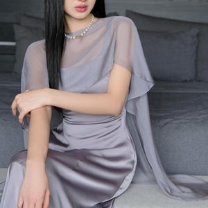 Pode incluir: Uma mulher vestindo um vestido de cetim cinza com uma sobreposi&ccedil;&atilde;o transparente e mangas de capa. Um colar de p&eacute;rolas adorna o pesco&ccedil;o. O vestido tem um corpete ajustado e uma saia fluida. A mulher tem cabelo longo e preto.