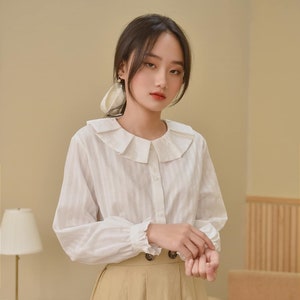 May include: A white long-sleeved blouse with a ruffled collar and button fastening. The blouse has thin white stripes and the sleeves have ruffles at the cuffs. The model is wearing a beige skirt.
