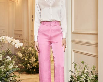 May include: A woman wearing a white button-down shirt and high-waisted, wide-leg pink pants. The outfit is paired with gold heels. The background features floral arrangements and a well-lit interior, suggesting a fashion or lifestyle theme.