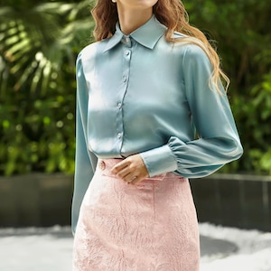 Pode incluir: Uma mulher usando uma camisa azul clara abotoada e uma saia rosa floral. A camisa tem mangas compridas e gola. Ela usa óculos de sol brancos e brincos pequenos. O fundo é verde desfocado.