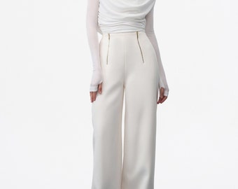 May include: A white draped top with long sleeves and shoulder pads, worn with high-waisted, wide-leg white trousers. The trousers have gold zips. The outfit is a monochromatic ensemble, suitable for formal occasions.