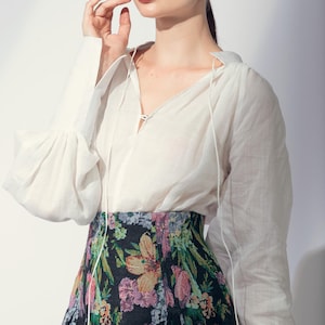 May include: A woman wearing a white blouse with bell sleeves and a V-neck. The blouse is paired with a floral print skirt featuring a variety of colors. The woman is also wearing gold earrings.