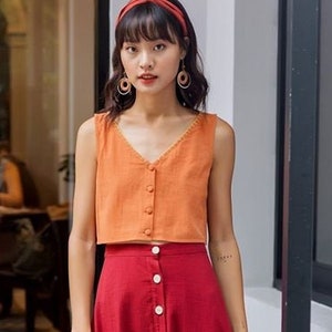 May include: An orange sleeveless top with a V-neck and button closure paired with a long, red button-down skirt. A small, woven handbag is held in the hand. The outfit is completed with a red headband and hoop earrings.
