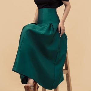 May include: A woman wearing a black off-the-shoulder top and a high-waisted, emerald green skirt. The skirt is pleated and falls to the ankles. She is seated on a wooden stool, wearing platform heels. The background is a solid beige.