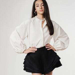 Puede incluir: Una mujer con una blusa blanca de manga larga y pantalones cortos negros. Los pantalones cortos tienen un diseño en capas. Lleva tacones blancos y un collar de perlas. El fondo es de color neutro con textura arenosa. El atuendo es elegante.