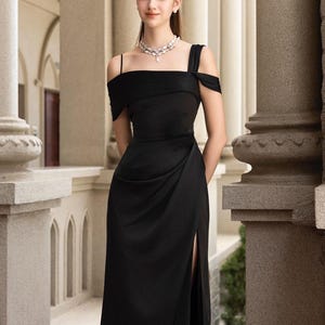 May include: A black midi dress with a side slit and off-the-shoulder sleeves. The dress is paired with silver sandals and a delicate necklace. The setting is an outdoor space with columns.
