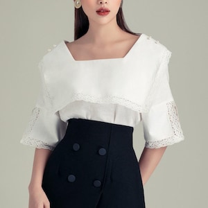 Puede incluir: Una blusa blanca con cuello cuadrado y ribete de encaje, combinada con una falda negra de cintura alta. La falda presenta un diseño envolvente y detalles de botones. El conjunto es elegante.