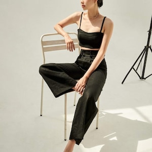 Puede incluir: Una mujer con un top negro corto y pantalones anchos se sienta en una silla de metal blanca. Lleva sandalias marrones y tiene el pelo recogido hacia atr&aacute;s.
