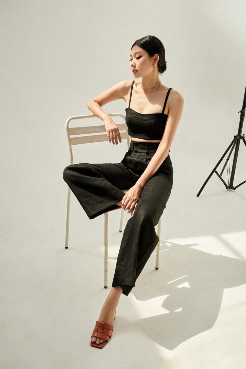 Puede incluir: Una mujer con un top negro corto y pantalones anchos se sienta en una silla de metal blanca. Lleva sandalias marrones y tiene el pelo recogido hacia atr&aacute;s.