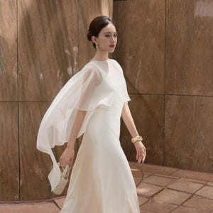Pode incluir: Um vestido de noiva branco com uma sobreposi&ccedil;&atilde;o de capa transparente. O vestido tem um design simples e elegante com uma saia fluida. A modelo est&aacute; usando uma pulseira de ouro e segurando uma pequena bolsa adornada.