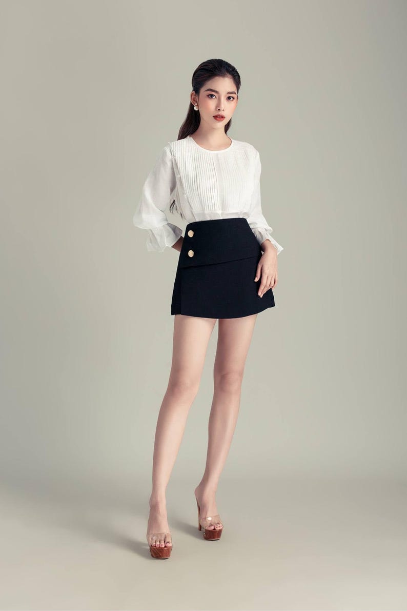 May include: A white blouse with pleated detailing and long sleeves, paired with a black mini skirt with gold buttons. The outfit is completed with platform heels. The outfit is worn by a person.