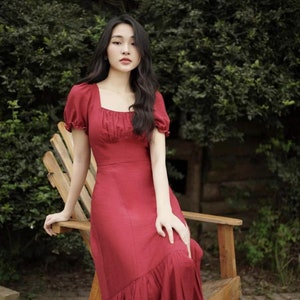 Puede incluir: Una mujer lleva un vestido midi rojo con escote cuadrado y mangas abullonadas. El vestido tiene un dobladillo con volantes y está hecho de una tela ligera. Está sentada en una silla de madera en un jardín.