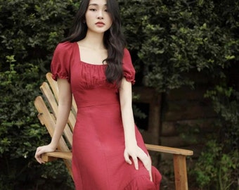 May include: A woman wearing a red midi dress with a square neckline and puff sleeves. The dress has a ruffled hem and is made of a lightweight fabric. She is sitting on a wooden chair in a garden setting.