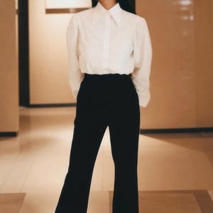 Puede incluir: Una mujer con una camisa blanca con mangas abullonadas y pantalones negros de cintura alta. El atuendo es de estilo cl&aacute;sico y minimalista, adecuado para diversas ocasiones. Los pantalones tienen un dise&ntilde;o de pierna ancha.