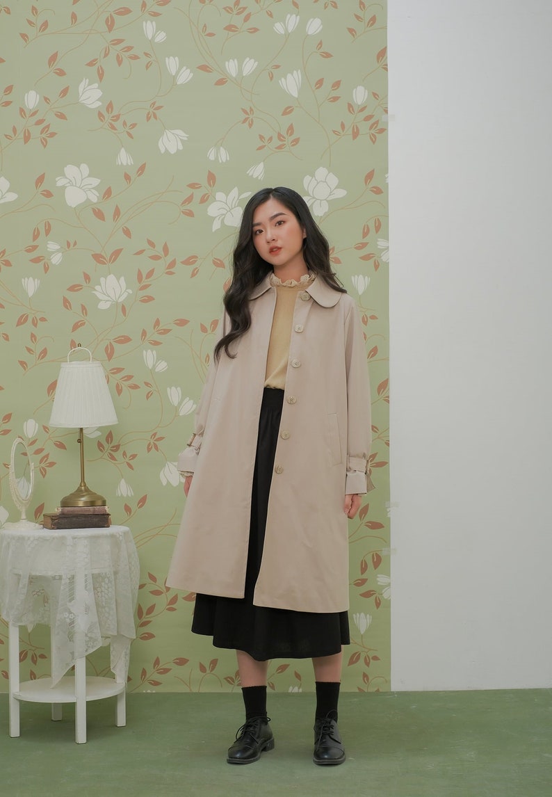 K&ouml;nnte beinhalten: Ein beiger Trenchcoat mit Knopfleiste und Peter-Pan-Kragen. Der Mantel hat lange &Auml;rmel mit dekorativen Kn&ouml;pfen an den Manschetten. Das Model tr&auml;gt einen schwarzen Rock und schwarze Schuhe.
