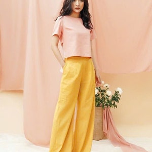 May include: A woman wearing a pink cropped top and wide-legged yellow pants. She is standing in front of a pink backdrop.