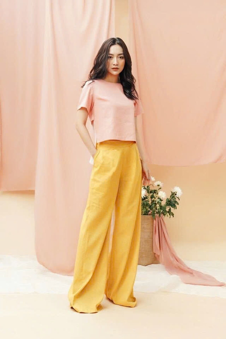 May include: A woman wearing a pink cropped top and wide-legged yellow pants. She is standing in front of a pink backdrop.