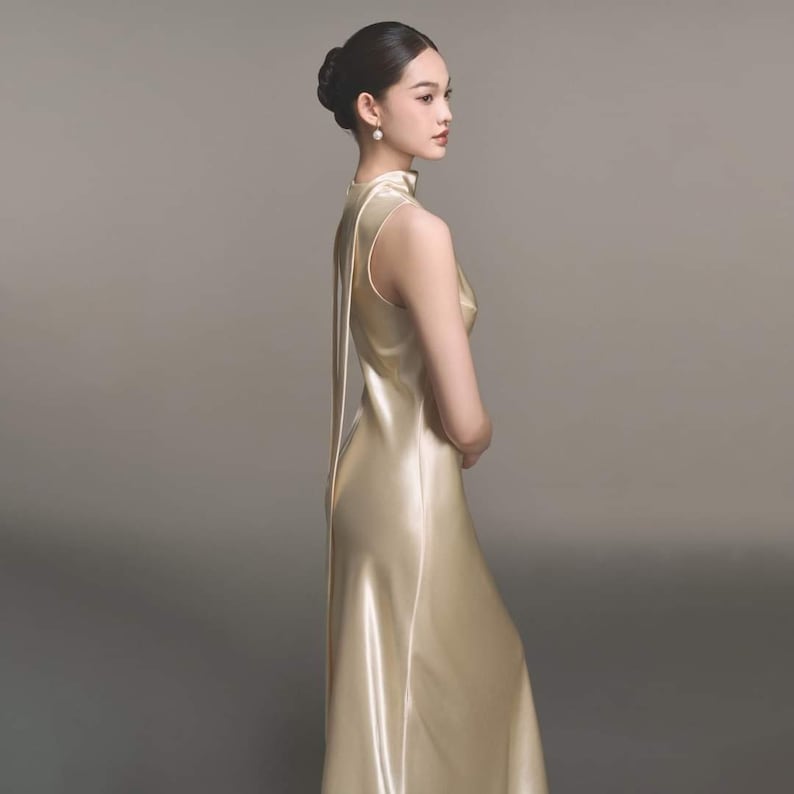 Puede incluir: Una mujer con un vestido de sat&eacute;n dorado sin mangas, cuello alto y cola larga. El vestido tiene un dise&ntilde;o simple y elegante.