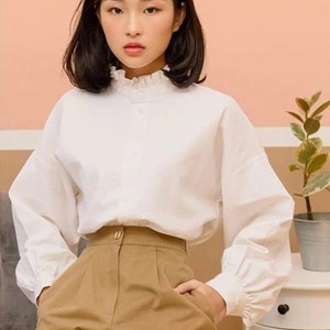 Puede incluir: Una blusa blanca de cuello con cuello fruncido y mangas largas. La blusa está metida en unos pantalones marrones de cintura alta.