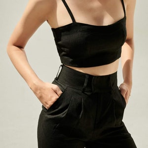 Puede incluir: Un top corto de lino negro con tirantes finos y un pantal&oacute;n de lino negro de cintura alta con pliegues. El atuendo lo lleva una persona con un collar de cadena dorada.