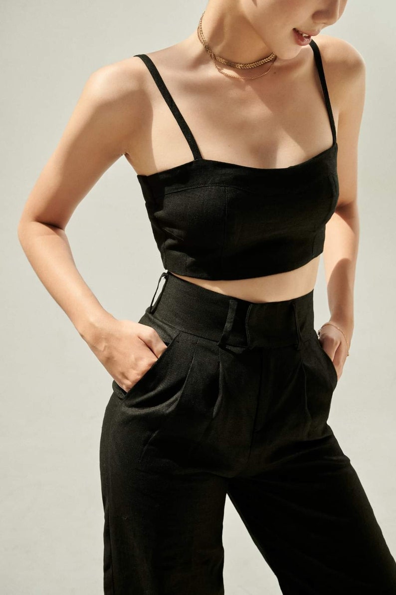 Puede incluir: Un top corto de lino negro con tirantes finos y un pantal&oacute;n de lino negro de cintura alta con pliegues. El atuendo lo lleva una persona con un collar de cadena dorada.