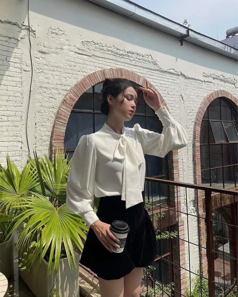 May include: A white blouse with a bow tie and long sleeves, worn with a black skirt. The blouse has a soft, flowing fabric, and the skirt has a velvet texture. A coffee cup is held in the hand.
