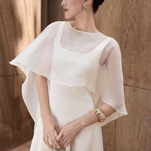 Pode incluir: Um vestido branco com uma sobreposi&ccedil;&atilde;o transparente em forma de capa. O vestido tem um decote quadrado e &eacute; adornado com uma pulseira e brincos dourados. O conjunto &eacute; elegante, ideal para eventos formais.