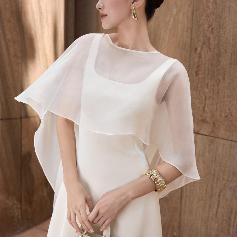 Pode incluir: Um vestido branco com uma sobreposi&ccedil;&atilde;o transparente em forma de capa. O vestido tem um decote quadrado e &eacute; adornado com uma pulseira e brincos dourados. O conjunto &eacute; elegante, ideal para eventos formais.