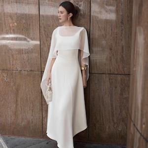 Pode incluir: Um vestido midi branco com decote quadrado e uma sobreposi&ccedil;&atilde;o transparente em forma de capa. O vestido tem uma silhueta sutil em linha A e &eacute; combinado com uma pequena bolsa creme. O fundo &eacute; uma parede marrom texturizada.