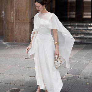 Pode incluir: Um vestido branco longo com uma sobreposi&ccedil;&atilde;o transparente em forma de capa. O vestido apresenta um corpete justo e uma saia fluida. Uma pequena bolsa branca e saltos a condizer completam o look. O conjunto &eacute; complementado com uma pulseira e brincos dourados.
