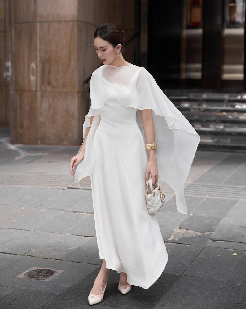 Pode incluir: Um vestido branco longo com uma sobreposi&ccedil;&atilde;o transparente em forma de capa. O vestido apresenta um corpete justo e uma saia fluida. Uma pequena bolsa branca e saltos a condizer completam o look. O conjunto &eacute; complementado com uma pulseira e brincos dourados.