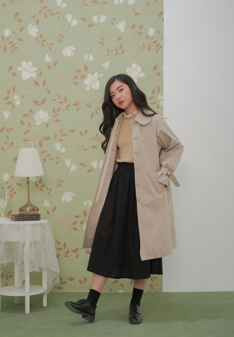 K&ouml;nnte beinhalten: Ein beiger Trenchcoat mit einem wei&szlig;en Streifen an den &Auml;rmeln und einem schwarzen Rock. Der Mantel hat einen Kragen mit wei&szlig;em Besatz und einen G&uuml;rtel in der Taille. Das Model tr&auml;gt schwarze Socken und schwarze Schuhe.