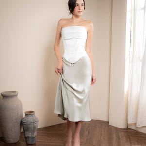 May include: A woman wearing a white strapless corset top and a light green satin midi skirt. The outfit is paired with patterned heels. The background includes a wooden floor, neutral walls, and a window with sheer curtains.