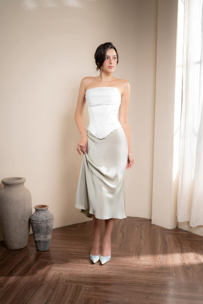 May include: A woman wearing a white strapless corset top and a light green satin midi skirt. The outfit is paired with patterned heels. The background includes a wooden floor, neutral walls, and a window with sheer curtains.