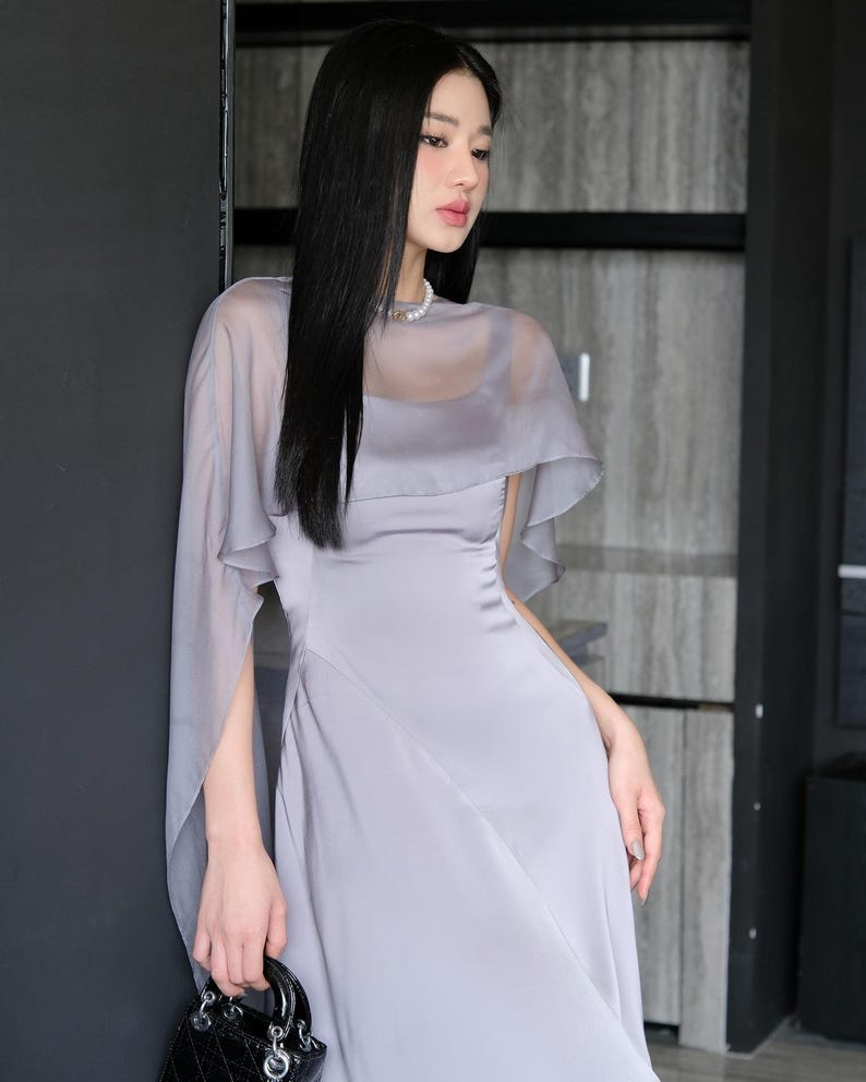 Pode incluir: Um vestido cinza claro com uma sobreposi&ccedil;&atilde;o de capa transparente. O vestido tem um corpete justo e uma saia fluida. Uma bolsa preta com uma al&ccedil;a superior &eacute; segurada. A modelo usa um colar de p&eacute;rolas.