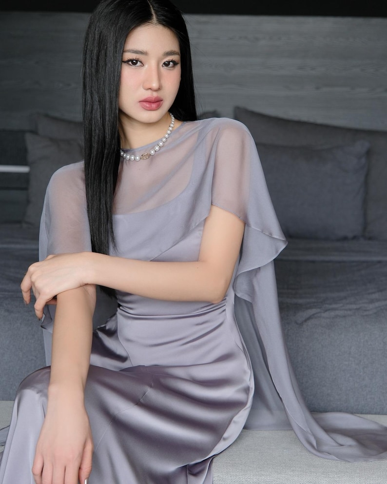 Pode incluir: Uma mulher vestindo um vestido de cetim cinza com uma sobreposi&ccedil;&atilde;o transparente e mangas de capa. Um colar de p&eacute;rolas adorna o pesco&ccedil;o. O vestido tem um corpete ajustado e uma saia fluida. A mulher tem cabelo longo e preto.