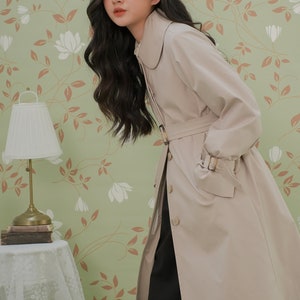 K&ouml;nnte beinhalten: Ein beiger Trenchcoat mit einem gro&szlig;en Kragen, einem G&uuml;rtel und langen &Auml;rmeln. Der Mantel wird von einer Person mit langen dunklen Haaren vor einem gr&uuml;nen floralen Hintergrund getragen.