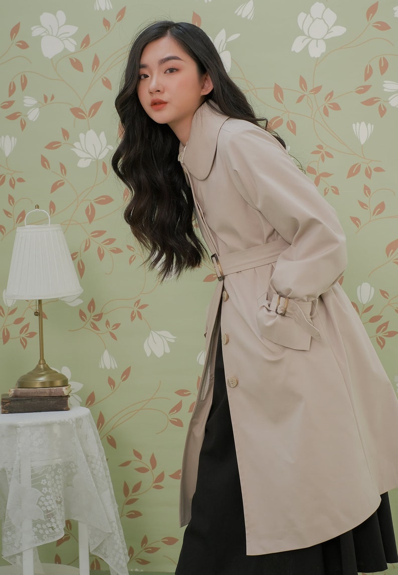 K&ouml;nnte beinhalten: Ein beiger Trenchcoat mit einem gro&szlig;en Kragen, einem G&uuml;rtel und langen &Auml;rmeln. Der Mantel wird von einer Person mit langen dunklen Haaren vor einem gr&uuml;nen floralen Hintergrund getragen.