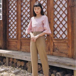 Pode incluir: Uma mulher vestindo uma blusa rosa com decote quadrado e cal&ccedil;as marrons est&aacute; em frente a uma porta de treli&ccedil;a de madeira. A porta tem um design asi&aacute;tico tradicional com um padr&atilde;o de cruz.