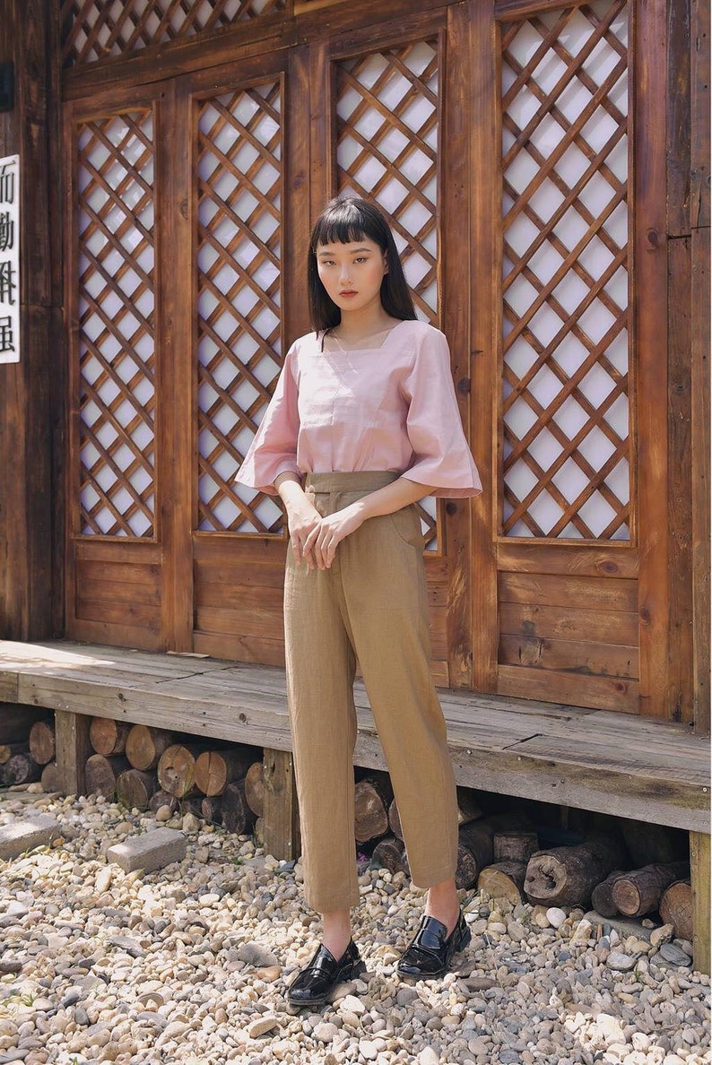 Pode incluir: Uma mulher vestindo uma blusa rosa com decote quadrado e cal&ccedil;as marrons est&aacute; em frente a uma porta de treli&ccedil;a de madeira. A porta tem um design asi&aacute;tico tradicional com um padr&atilde;o de cruz.
