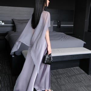 Pode incluir: Um conjunto fluido cinza claro com cal&ccedil;as de perna larga e um top a condizer em forma de capa. O traje &eacute; complementado com saltos pretos e uma pequena bolsa preta texturizada. O visual geral &eacute; elegante e sofisticado, adequado para ocasi&otilde;es formais.