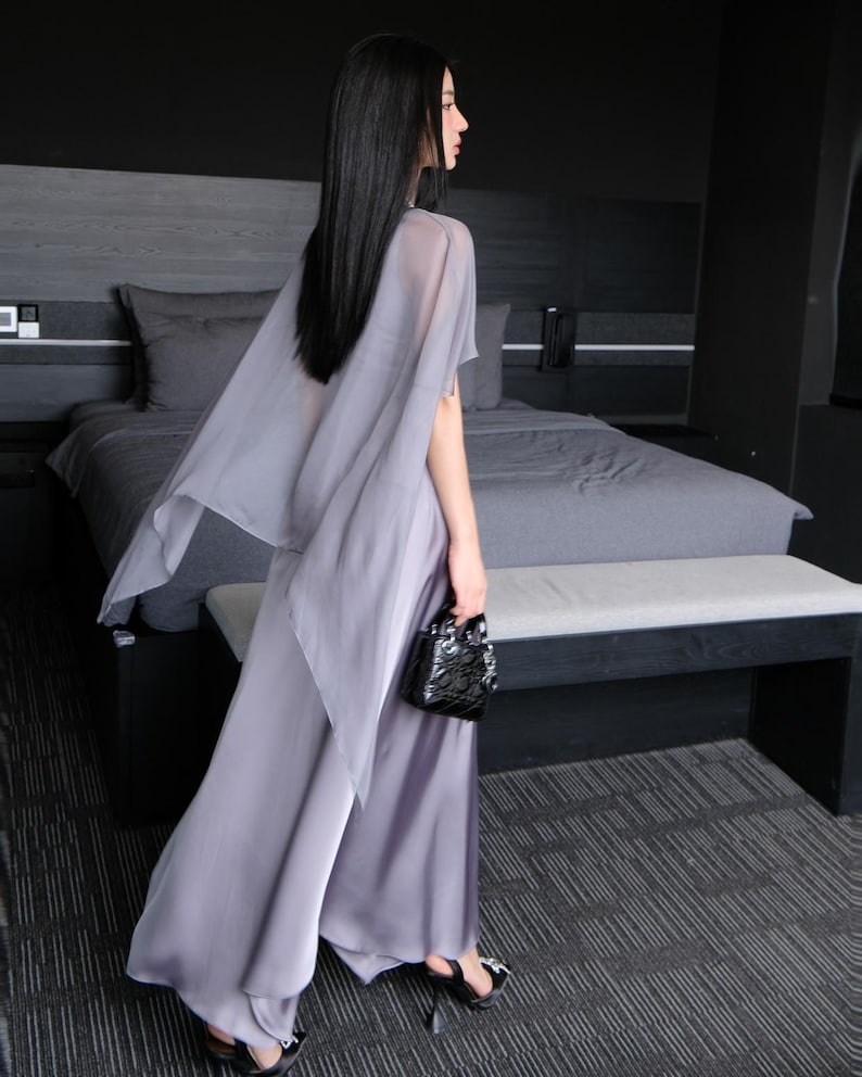 Pode incluir: Um conjunto fluido cinza claro com cal&ccedil;as de perna larga e um top a condizer em forma de capa. O traje &eacute; complementado com saltos pretos e uma pequena bolsa preta texturizada. O visual geral &eacute; elegante e sofisticado, adequado para ocasi&otilde;es formais.