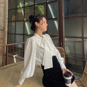 May include: A white blouse with a bow tie and long, puffed sleeves, worn with a black velvet skirt. The person is holding a coffee cup with a black lid. The setting is a cafe with a large window.