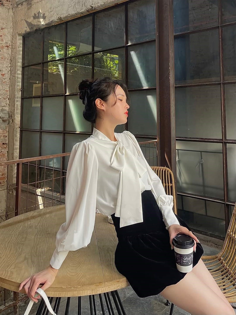 May include: A white blouse with a bow tie and long, puffed sleeves, worn with a black velvet skirt. The person is holding a coffee cup with a black lid. The setting is a cafe with a large window.