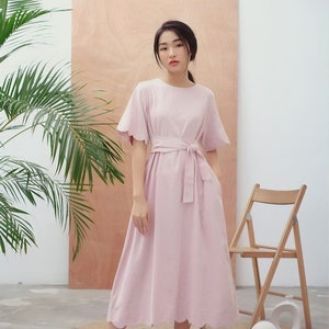 Puede incluir: Vestido rosa claro con dobladillo y mangas festoneadas. El vestido tiene un cinturón a juego atado a la cintura. La modelo lleva zapatos negros. Una silla de madera y una planta están en el fondo.