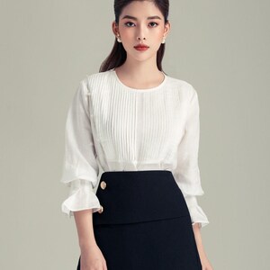 May include: A white blouse with pleated detailing and three-quarter sleeves, paired with a black mini skirt. The skirt features a layered design and gold-coloured buttons. The outfit is a classic and elegant ensemble.