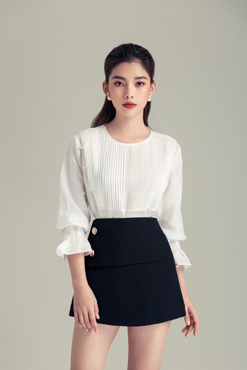 May include: A white blouse with pleated detailing and three-quarter sleeves, paired with a black mini skirt. The skirt features a layered design and gold-coloured buttons. The outfit is a classic and elegant ensemble.