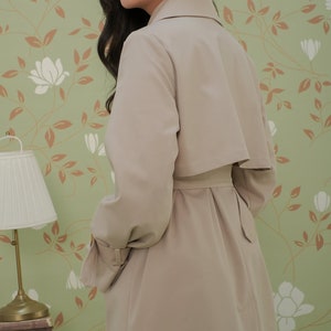 K&ouml;nnte beinhalten: Ein beiger Trenchcoat mit G&uuml;rtel und Kragen. Der Mantel ist aus glattem Stoff und hat ein klassisches Design.